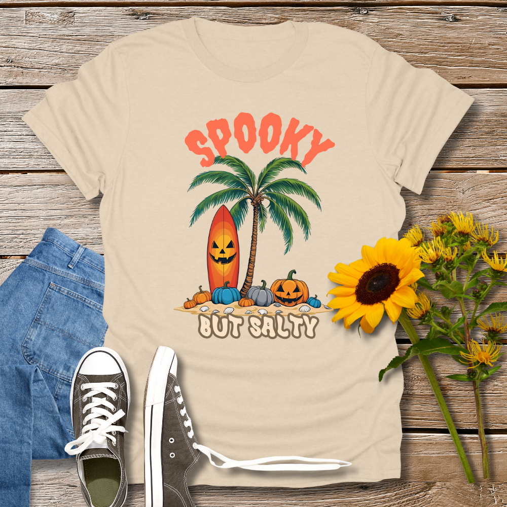 Beige t-shirt with Halloween-themed design featuring palm trees and pumpkins on a wooden surface with jeans and sneakers.
