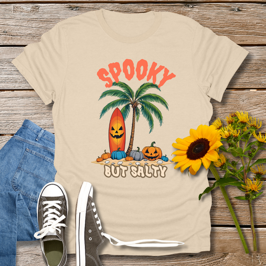 Beige t-shirt with Halloween-themed design featuring palm trees and pumpkins on a wooden surface with jeans and sneakers.