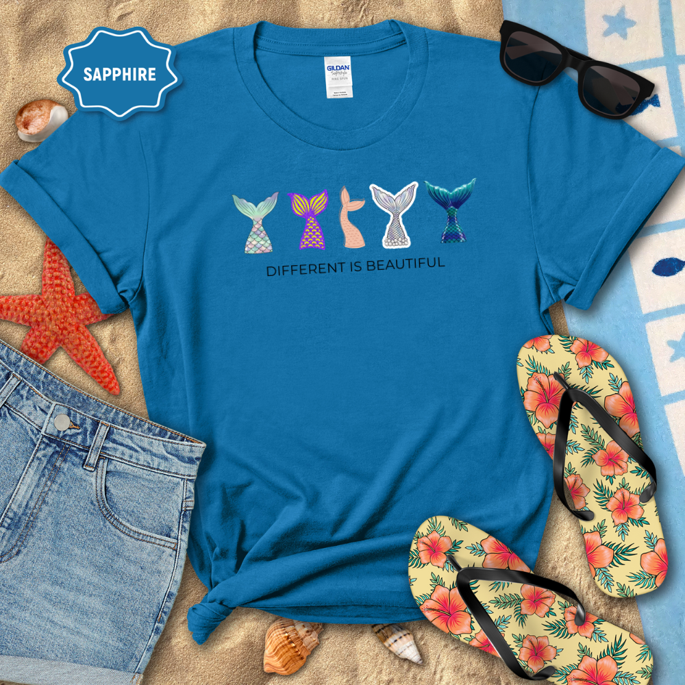 Blue t-shirt with colorful mermaid tail designs and 'Different is Beautiful' text, surrounded by beach accessories.