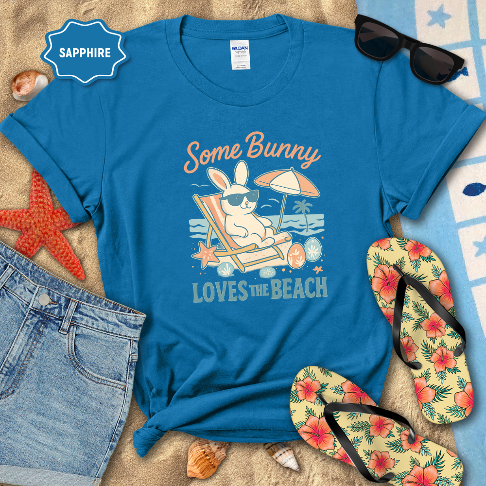 Blue t-shirt with beach-themed design featuring a bunny, sunglasses, and flip-flops on sand.