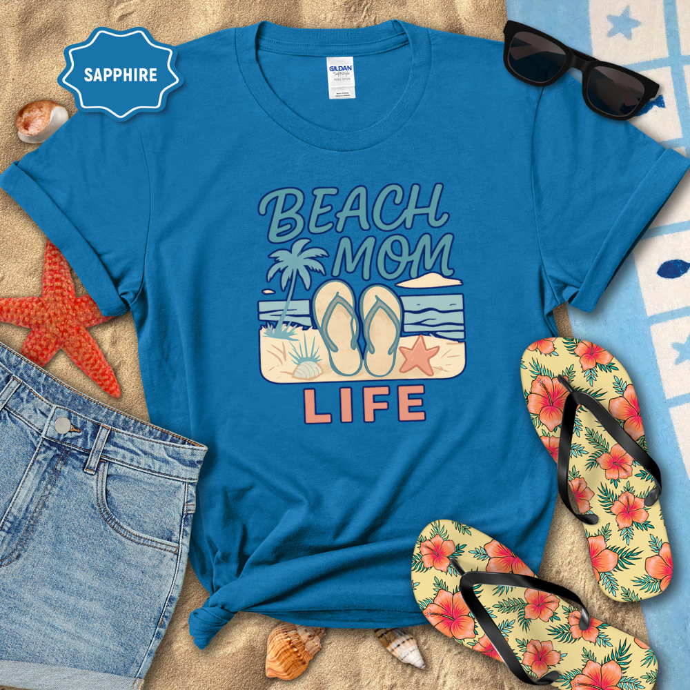 Blue t-shirt with 'Beach Mom Life' design on sand with sunglasses, shorts, and flip-flops.