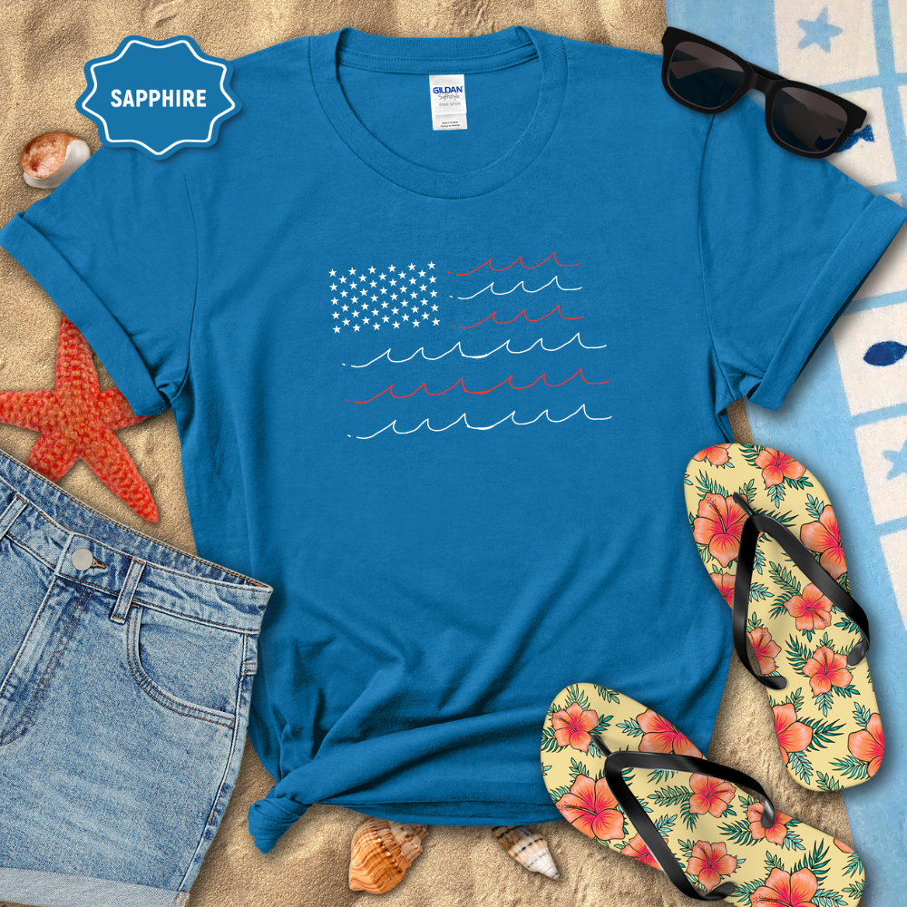 Blue t-shirt with American flag design on a sandy surface with sunglasses, flip-flops, and denim shorts.