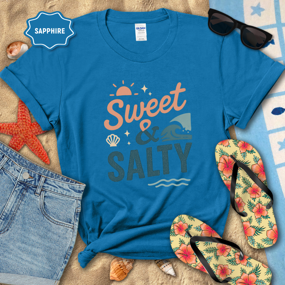 Blue t-shirt with 'Sweet & Salty' design on a sandy surface with beach accessories.