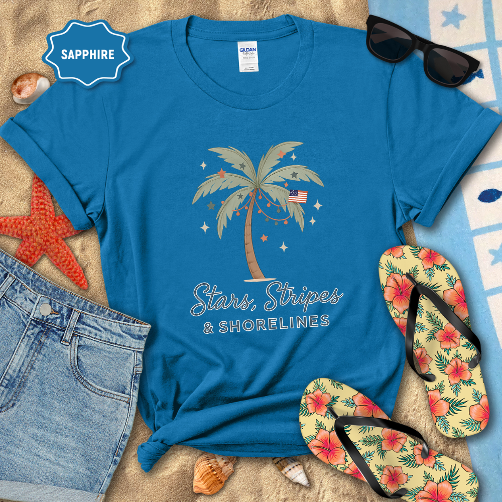 Blue t-shirt with palm tree design and text, surrounded by beach accessories on sand.