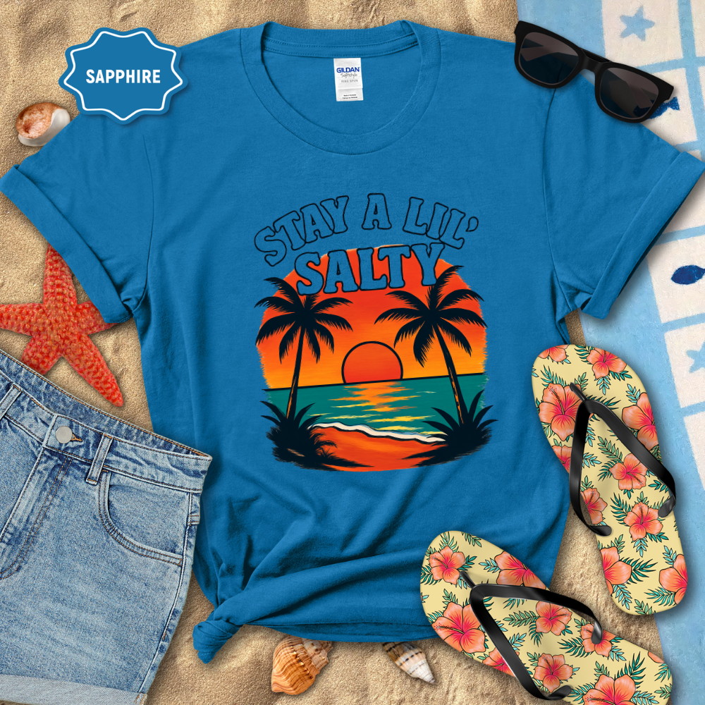 Blue t-shirt with 'Stay a Lil' Salty' design on a beach-themed background with sunglasses, flip-flops, and denim shorts.