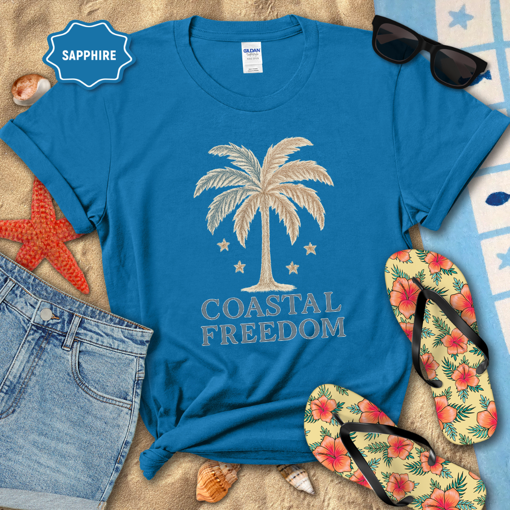 Blue t-shirt with palm tree design and 'Coastal Freedom' text, surrounded by beach accessories on sand.