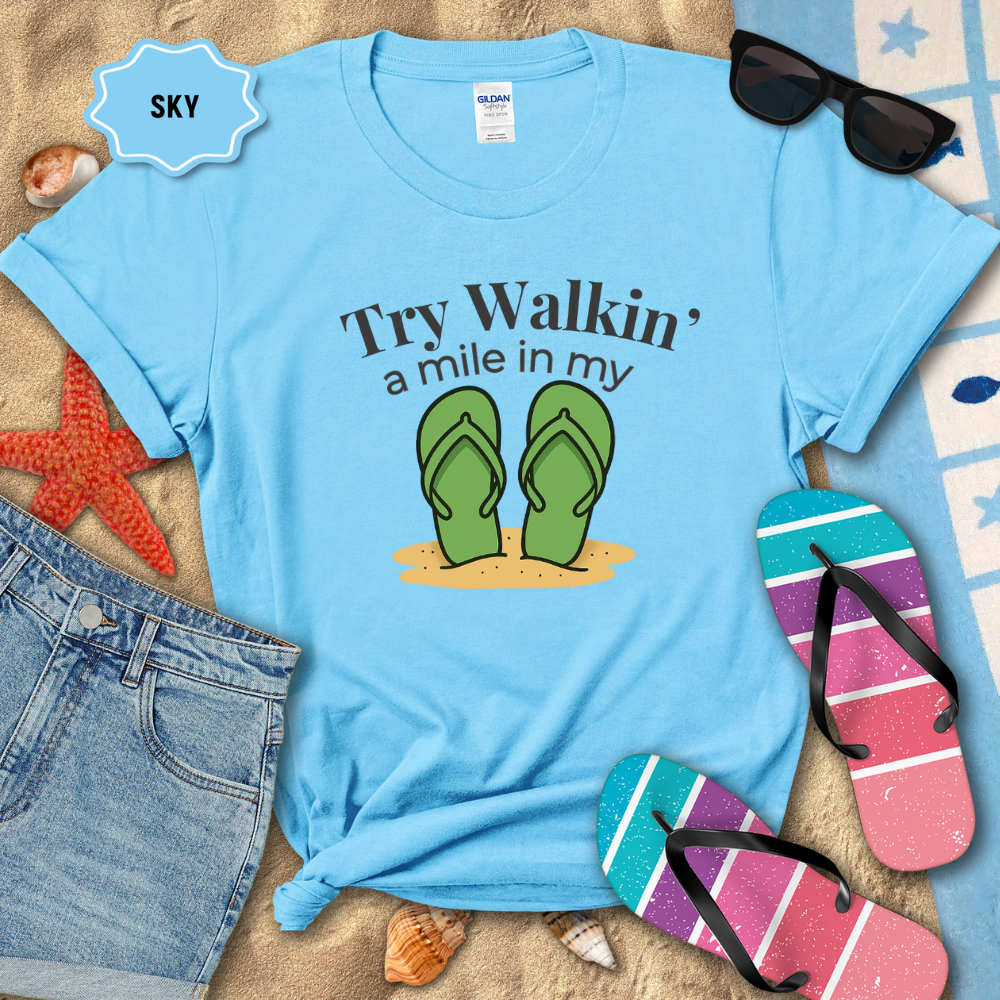 Blue t-shirt with flip-flop design and text on a sandy background with beach accessories.