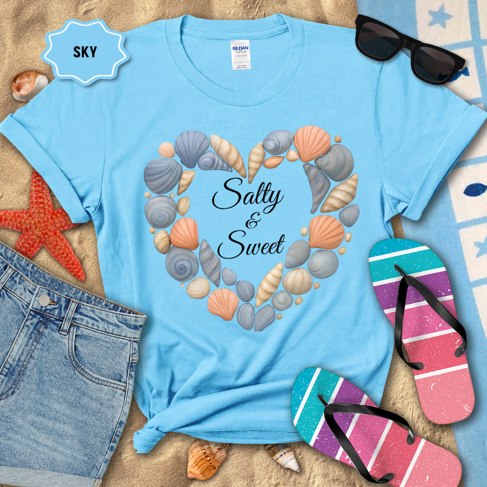 Light blue t-shirt with seashell design and 'Salty & Sweet' text, surrounded by beach accessories on sand.