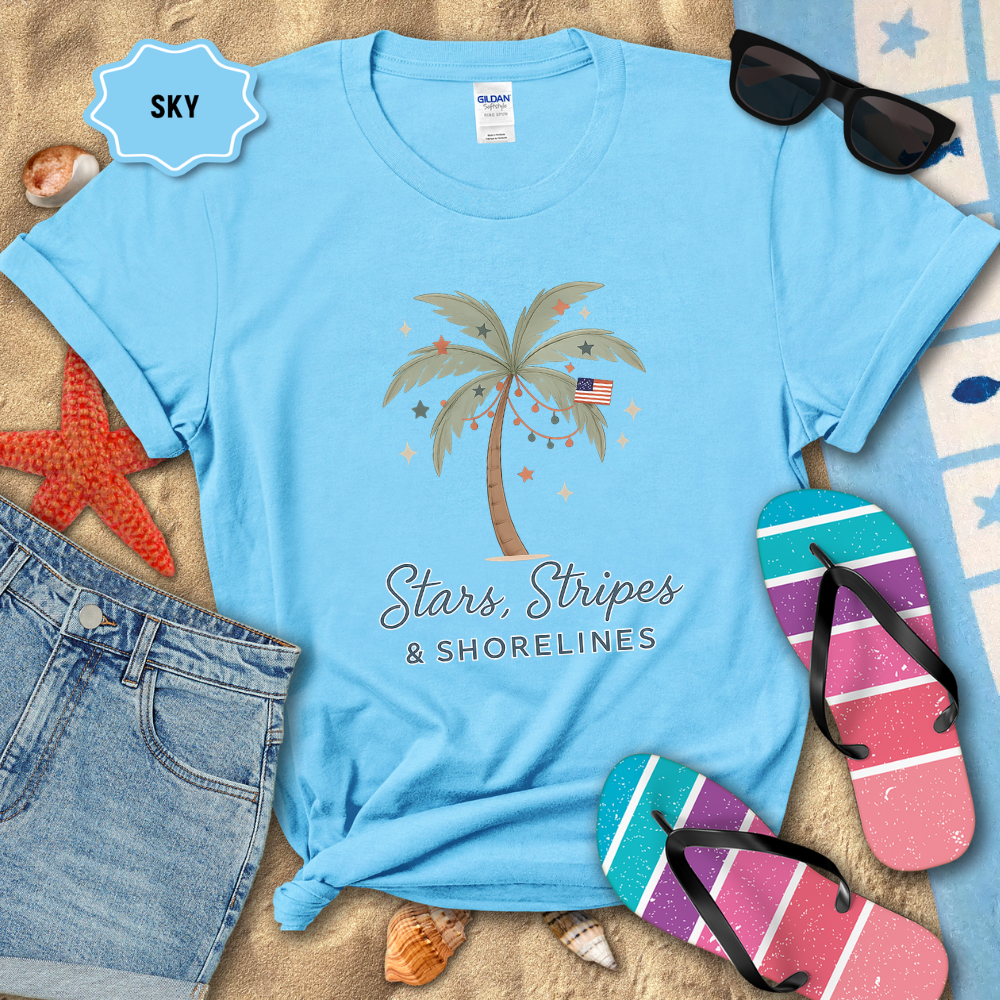 Blue t-shirt with palm tree design and text, surrounded by beach accessories on sand.