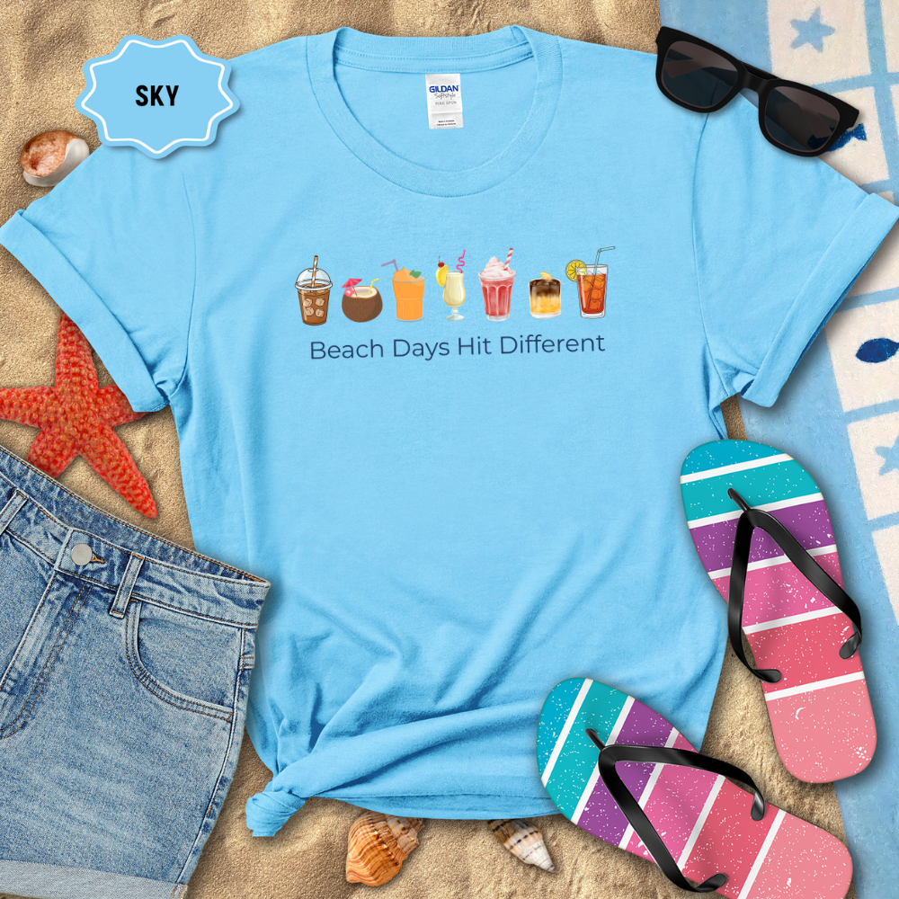 Light blue t-shirt with beach-themed graphics and text, surrounded by beach accessories on sand.