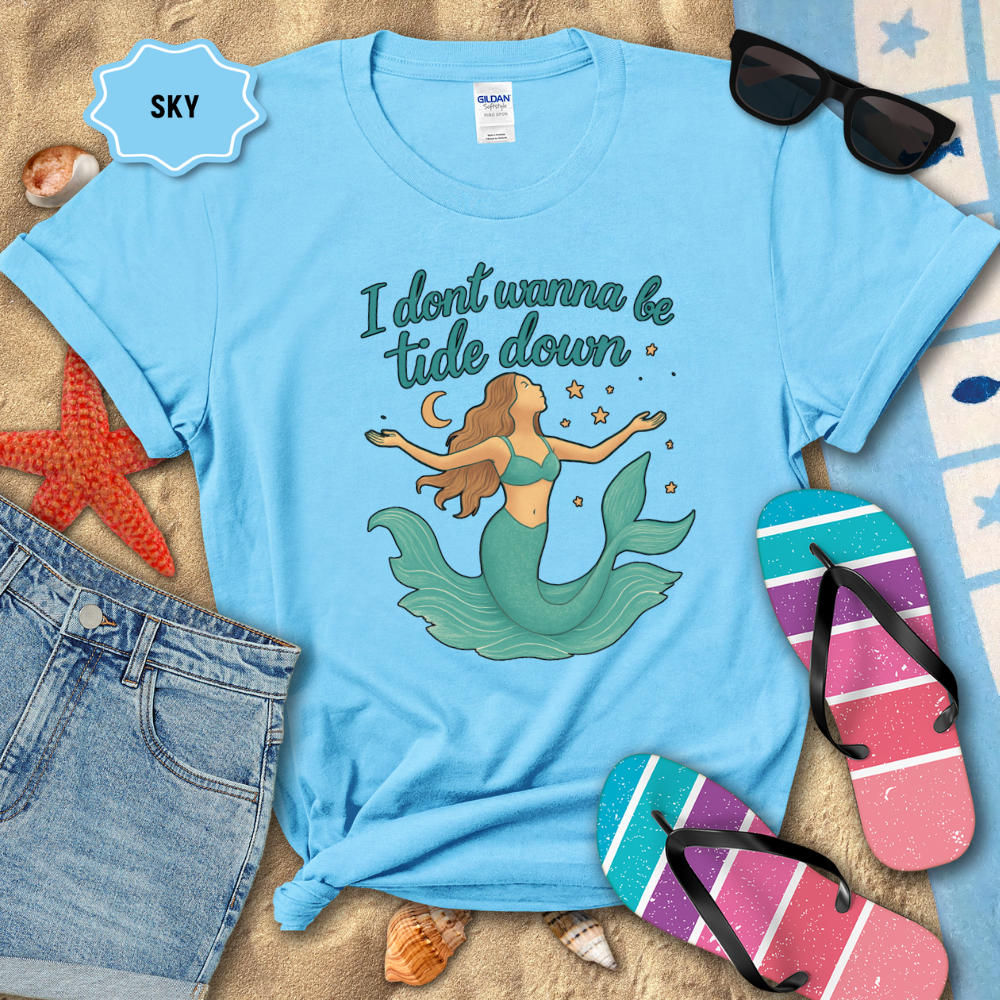 Light blue t-shirt with mermaid graphic and text, surrounded by beach items on sand.