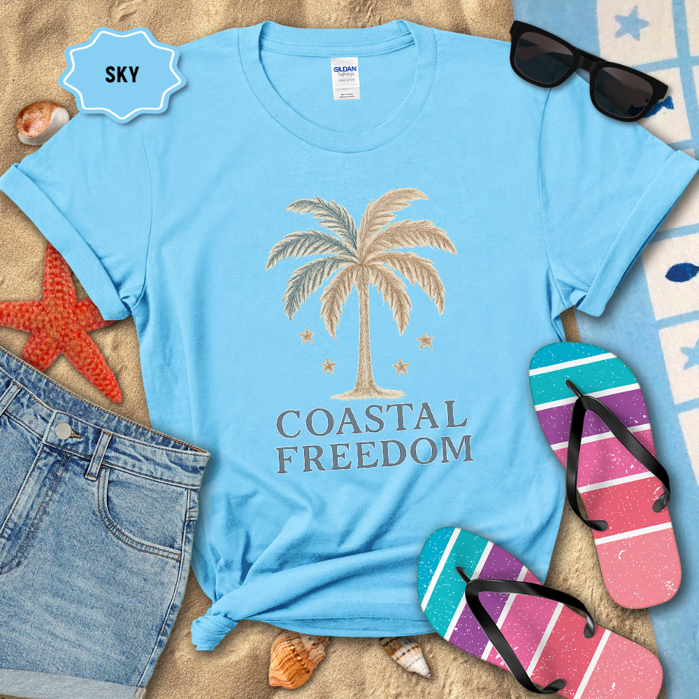 Blue t-shirt with palm tree design and 'Coastal Freedom' text on a sandy background with beach accessories.