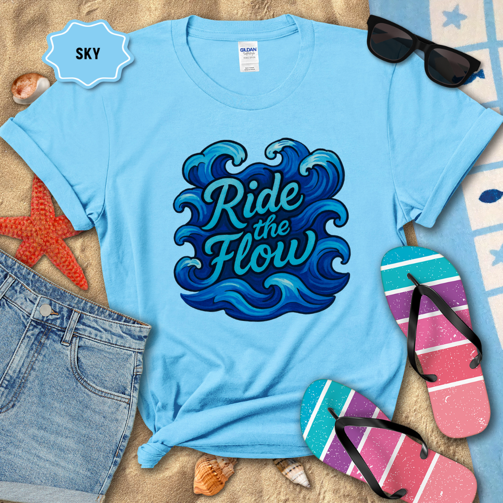 Light blue t-shirt with 'Ride the Flow' design on a sandy background with beach accessories.