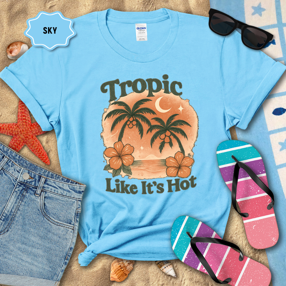 Blue t-shirt with tropical design and text, surrounded by beach accessories on sand.