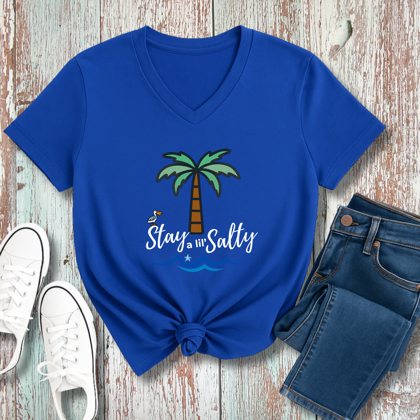 Blue t-shirt with palm tree design and 'Stay Salty' text on a wooden surface with jeans and sneakers.