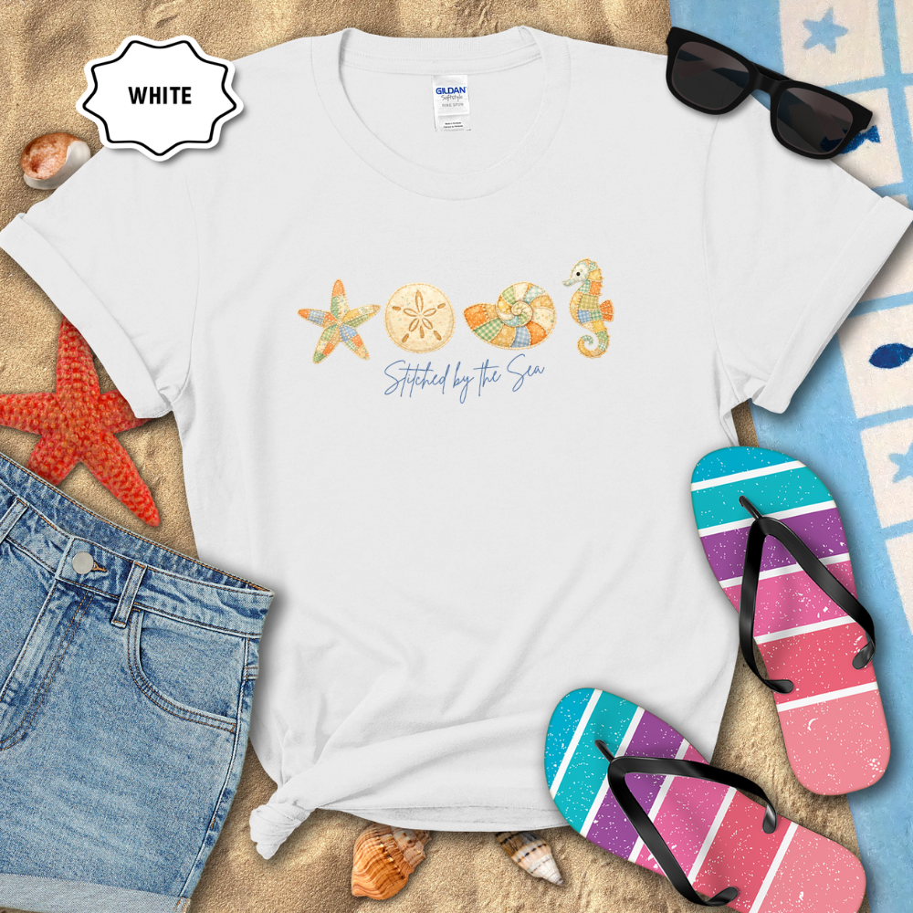 White t-shirt with beach-themed designs on a sandy surface with sunglasses, flip-flops, and denim shorts.
