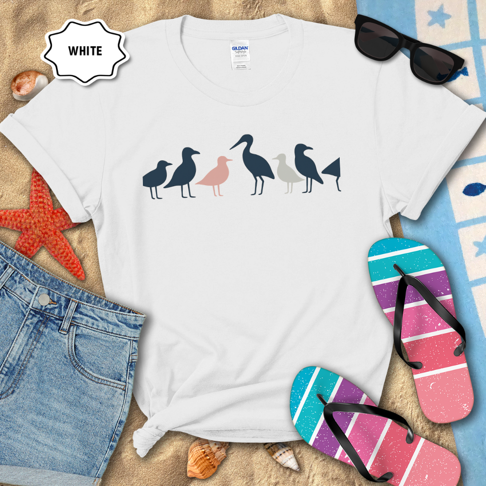 White t-shirt with bird design on a sandy surface with flip-flops and sunglasses.