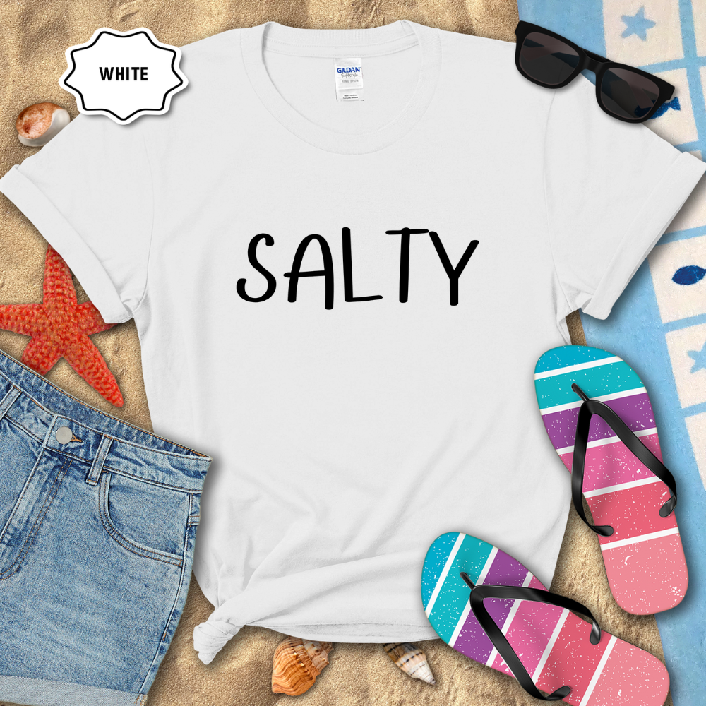 White t-shirt with a colorful turtle design on a sandy surface with beach accessories.