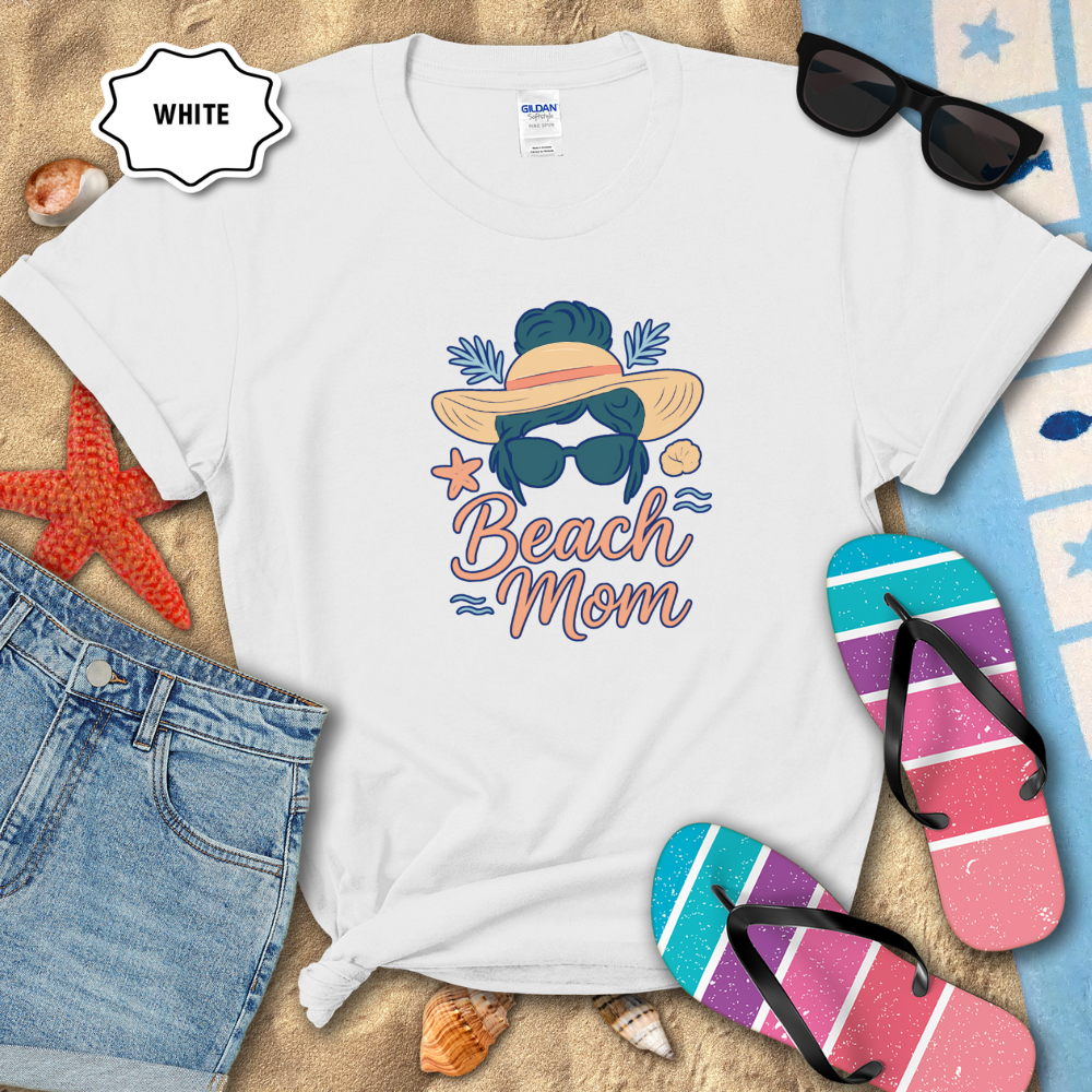 White t-shirt with 'Beach Mom' design on sand with sunglasses, flip-flops, and denim shorts.