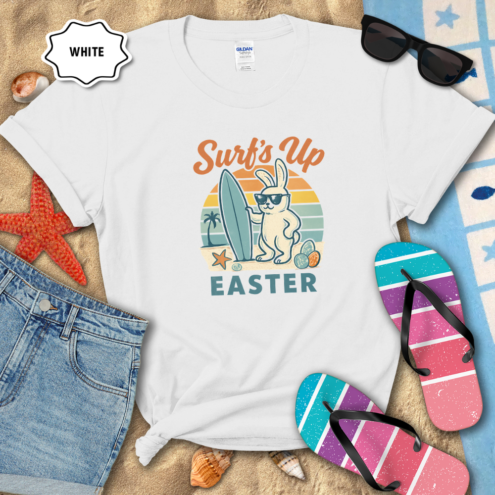 White t-shirt with 'Surf's Up Easter' design on a sandy background with flip-flops and sunglasses.