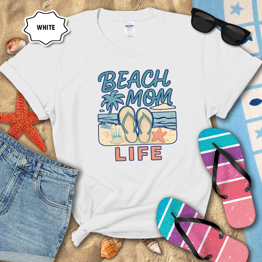 White t-shirt with 'Beach Mom Life' design on sand with flip-flops and sunglasses.