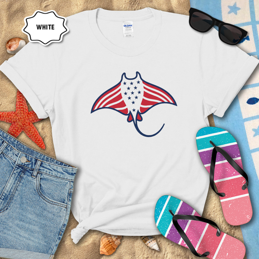 White t-shirt with a manta ray design featuring the American flag, surrounded by beach items on sand.