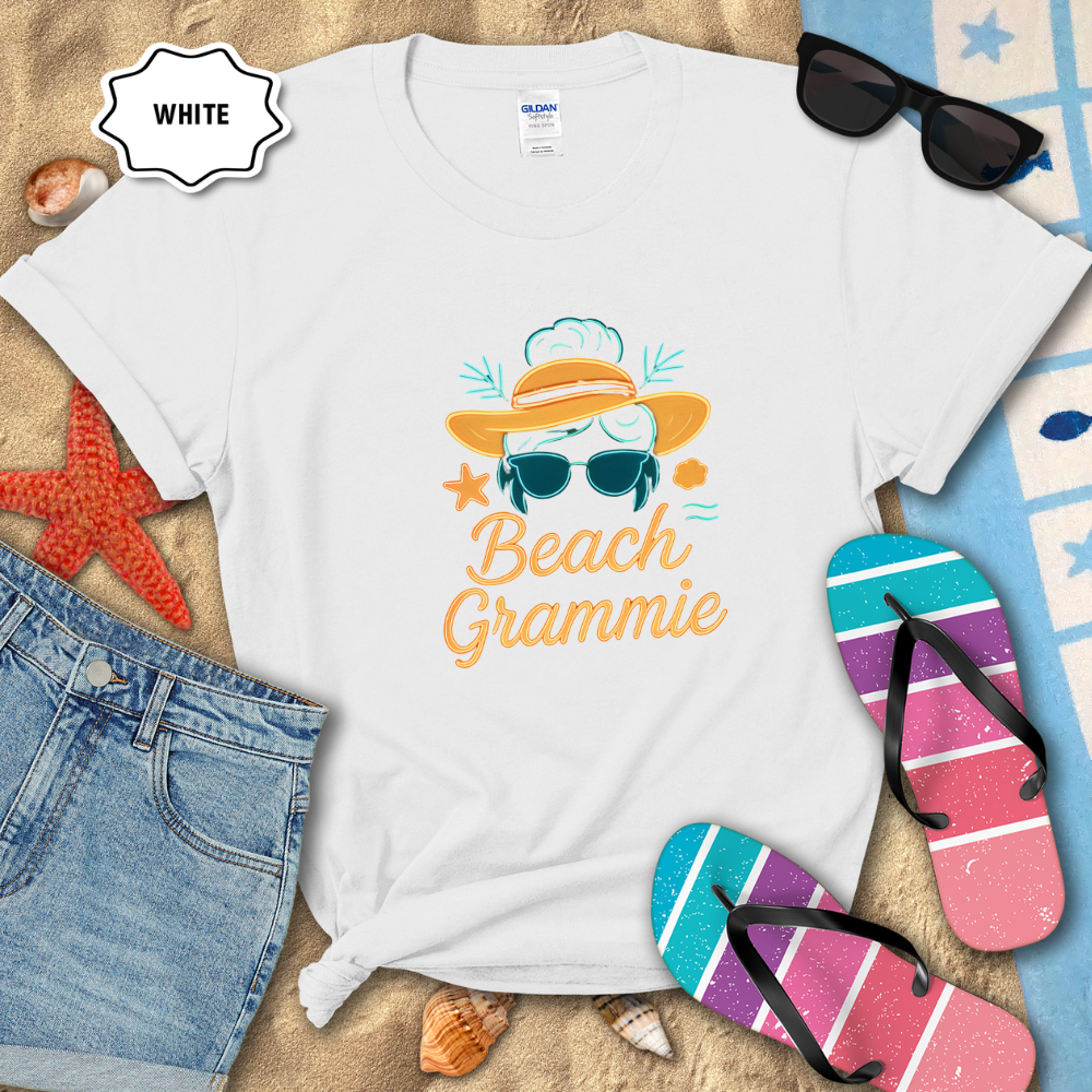 White t-shirt with 'Beach Grammie' design on sand with sunglasses, flip-flops, and denim shorts.