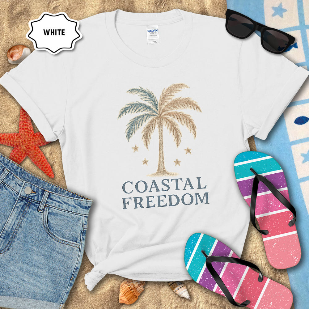 White t-shirt with palm tree design and 'Coastal Freedom' text, surrounded by beach accessories on sand.