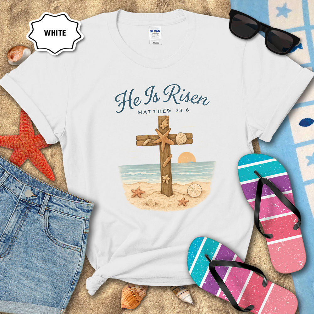 White t-shirt with 'He Is Risen' design on a sandy surface with beach accessories.