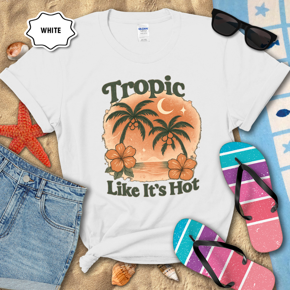 White t-shirt with tropical design and text, surrounded by beach accessories on sand.