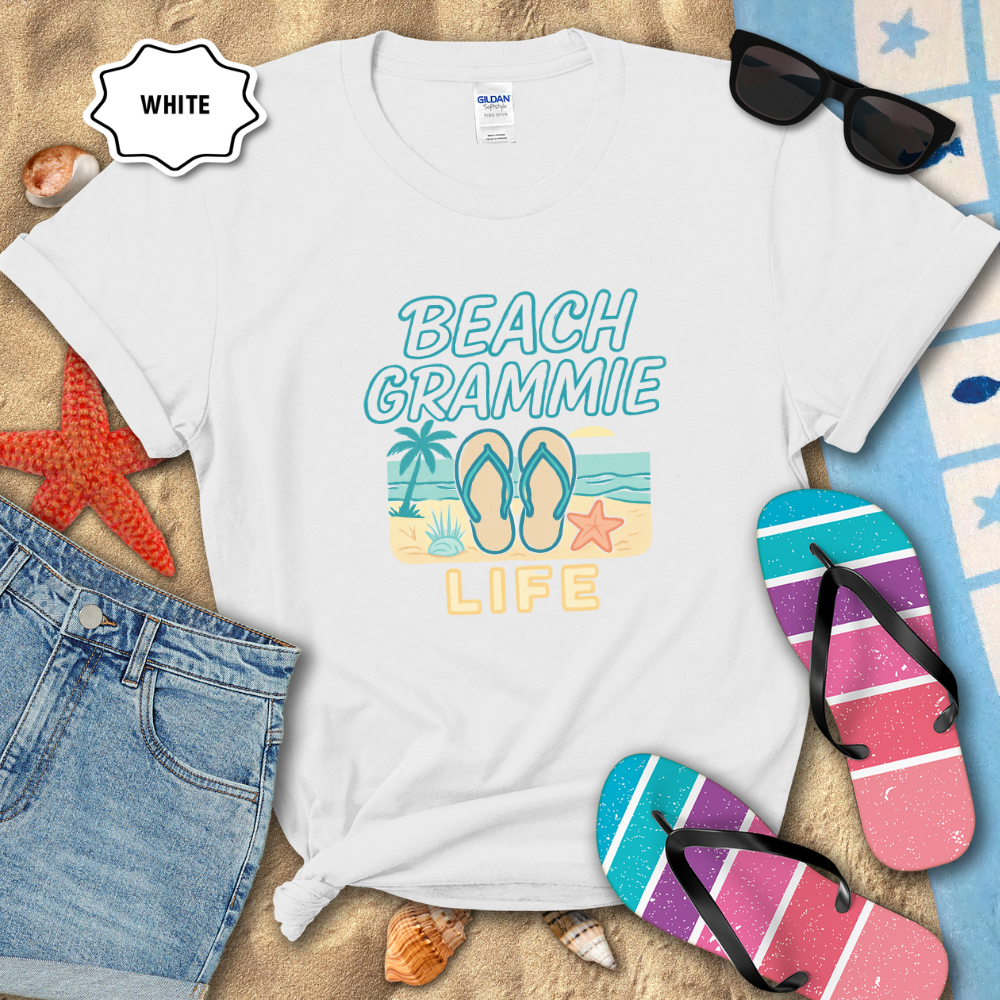 White t-shirt with 'Beach Grammie Life' design on sand with flip-flops and sunglasses.