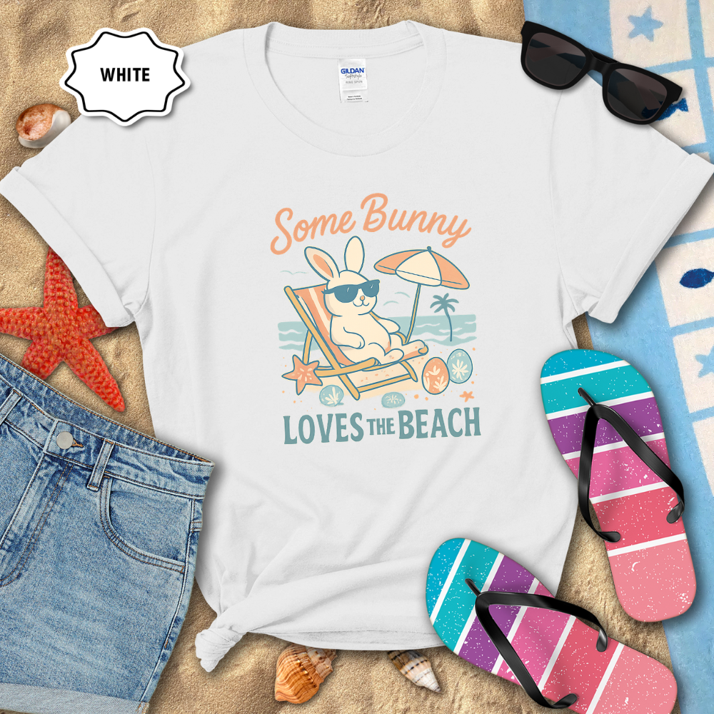 White t-shirt with bunny graphic and text 'Some Bunny Loves the Beach' on a sandy background with flip-flops and sunglasses.