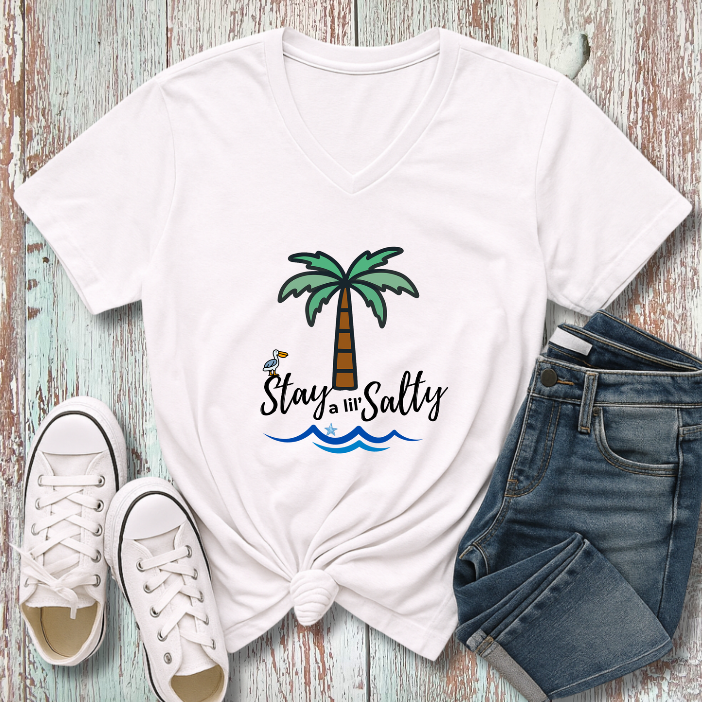 White t-shirt with palm tree and 'Stay Salty' text, paired with white sneakers and blue jeans on a wooden surface.