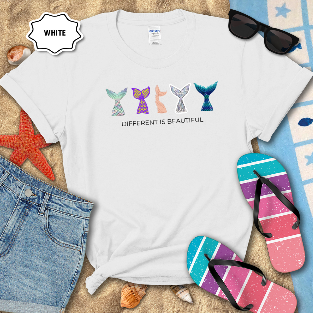 White t-shirt with mermaid tail designs and 'Different is Beautiful' text on a beach setting with sunglasses, flip-flops, and denim shorts.