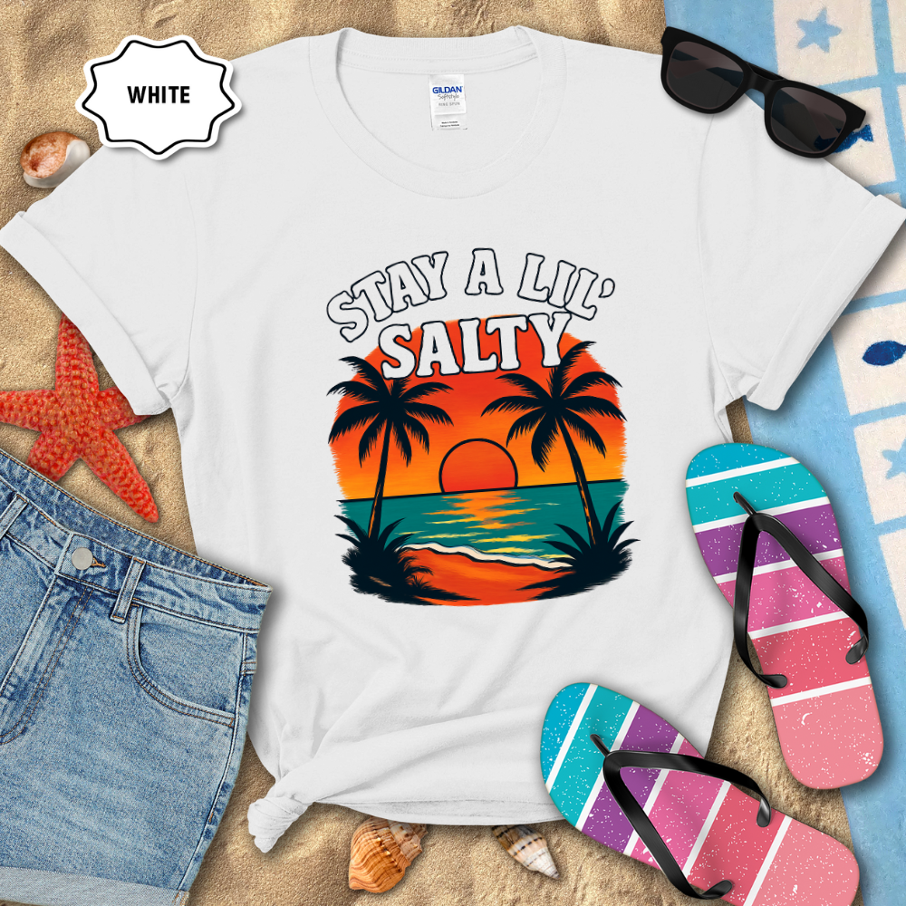 White t-shirt with 'Stay a Lil' Salty' design on a sandy background with flip-flops and sunglasses.