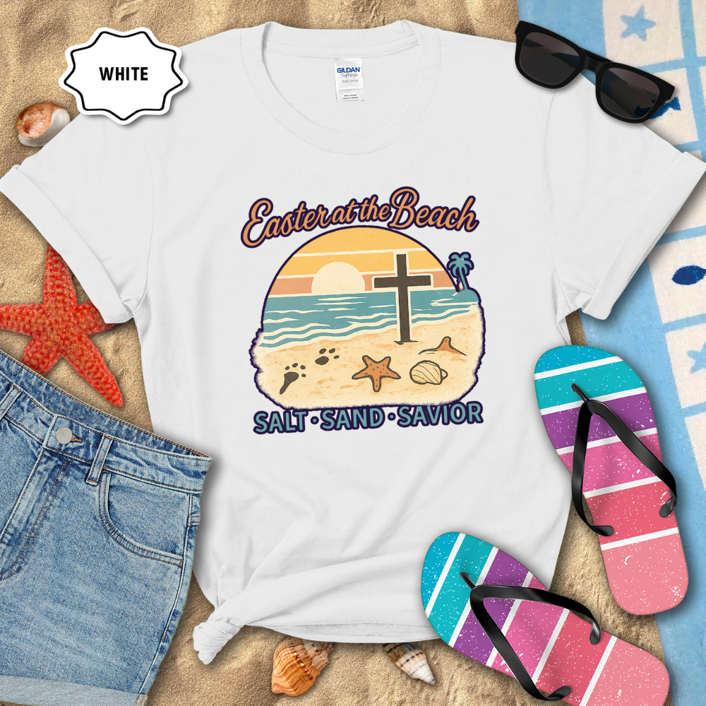 White t-shirt with beach-themed design on sand with sunglasses and flip-flops.