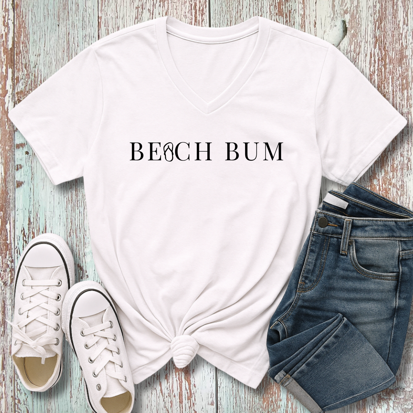 White t-shirt with 'BEACH BUM' text, white sneakers, and blue jeans on a wooden surface.