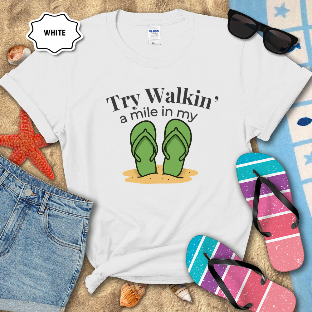White t-shirt with flip-flop graphic and text on a sandy background with beach accessories.