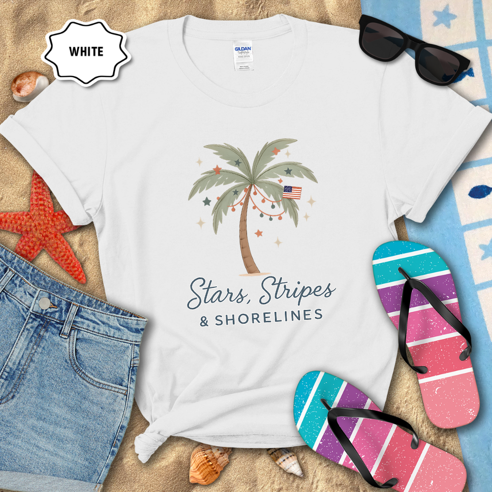 White t-shirt with palm tree design and 'Coastal Freedom' text, surrounded by beach accessories on sand.