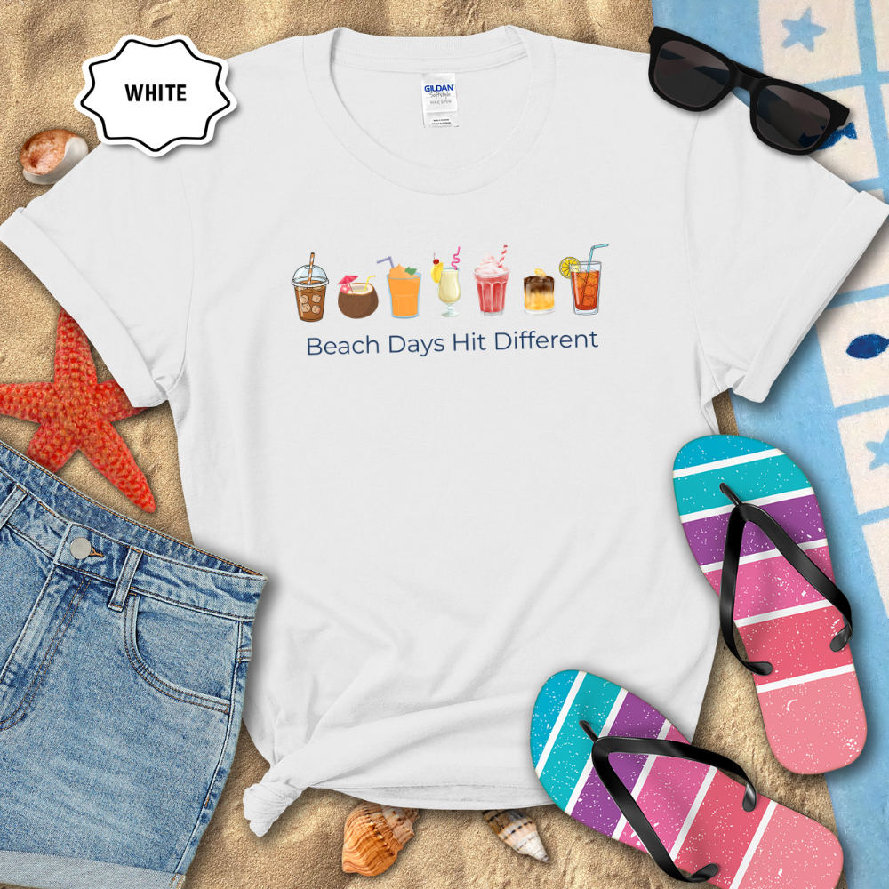 White t-shirt with beach-themed design and text, surrounded by flip-flops and sunglasses on a sandy background.