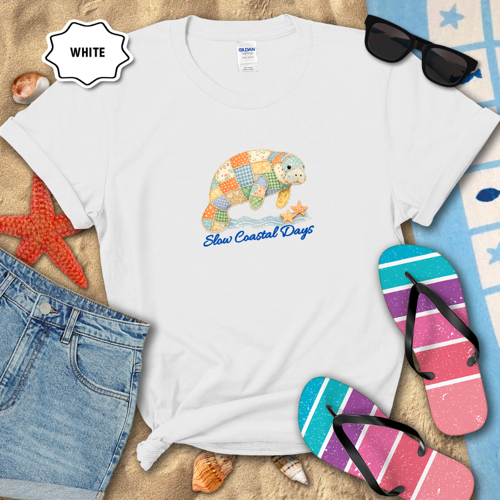 White t-shirt with a colorful turtle design and 'Sloppy Coastal Days' text, placed on sand with beach accessories.